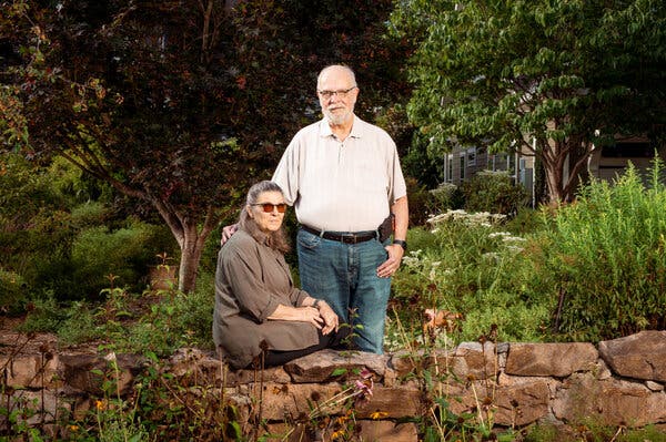 Joan Brasier sits on a stone wall while her husband, Paul Wilczynski, stands behind her, his hand on her upper back. Lots of trees and shrubbery are behind them.